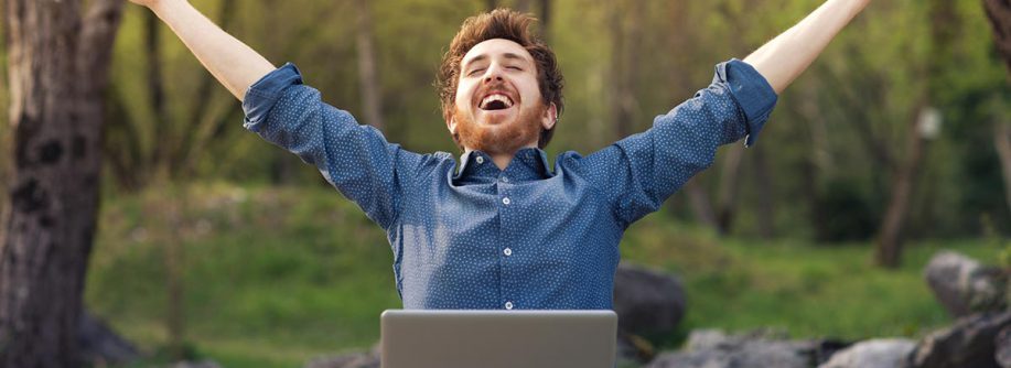 Ein Mann mit Laptop auf - Beruflicher Wechsel dem Schoß im Wald wirft glücklich die Arme hoch und freut sich über neuen Job