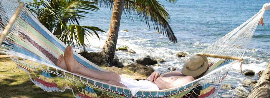 Ein Mann liegt entspannt in seiner Hängematte am Strand im Ausland unter Palmen, mit Hut auf dem Gesicht, und genießt sein Leben, weil er weiß, dass ein Qualitätsmakler sich um den Verkauf seiner Immobilie in Deutschland kümmert | Immobilienverkauf