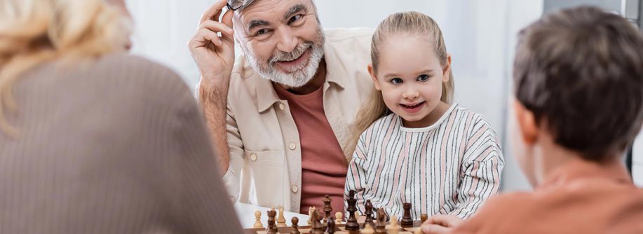 Ein Seniorenpärchen sitzt mit seinen Enkelkindern glücklich im Wohnzimmer und spielt Schach als Symbolbild für strategisch kluge Entscheidungen bei Investments für die Altersvorsorge | Wohnen im Alter