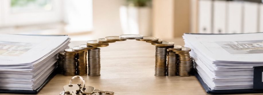 Münzstapel in Form eines Hauses zwischen Immobilienunterlagen und Schlüsseln symbolisieren eine Zwischenfinanzierung beim Immobilienkauf. I Zwischenfinanzierung